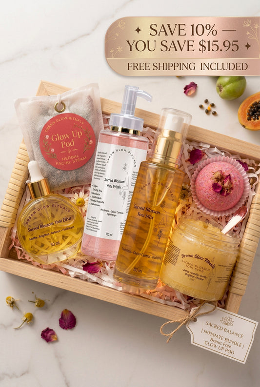 A curated self-care gift set arranged in a wooden tray with shredded paper filler, featuring a Glow Up Pod tea bag, a pink Sacred Balance body wash bottle, a golden Sacred Rituals oil bottle, a bath bomb, and a jar of body butter, with dried flowers and green fruits around it on a light marble surface.   