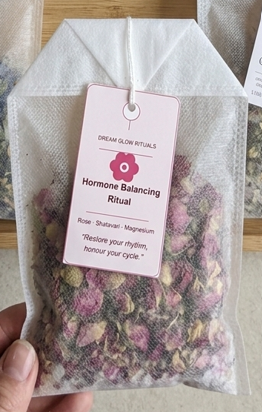 A hand holding a white mesh tea bag with a pink tag reading 'Hormone Balancing Ritual' from Dream Glow Rituals, filled with dried pink and green botanicals.
