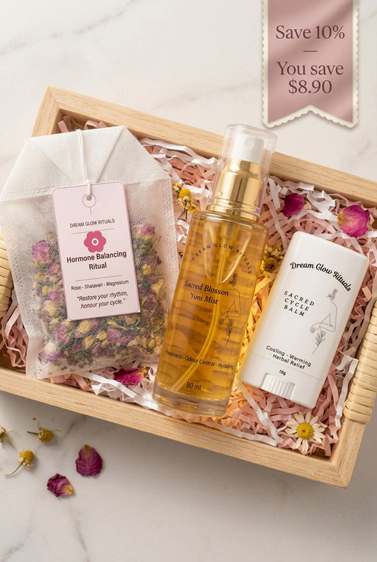 A skincare gift set from Dream Glow Skin Essentials arranged in a light wood tray with pink shredded paper and dried flower petals. The set includes an 'Hormone Balancing Ritual' tea bag, a 'Secret Weapon' toner mist in a clear bottle with a gold cap, and a 'Sacred Geometry' deodorant stick.