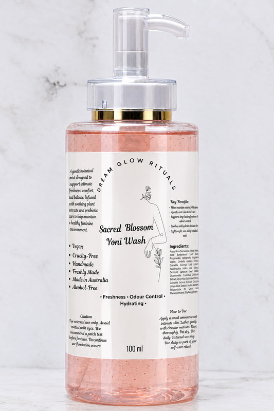 A 100ml bottle of Sacred Blossom Toni Wash by Dream Glow Rituals, featuring a clear pump dispenser with a gold collar and a white label, filled with pinkish-amber liquid, set against a light marble background.
